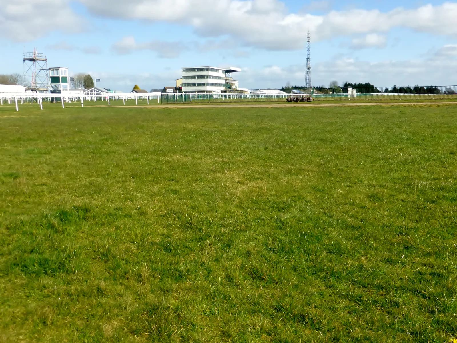 A wide, green field stretches out in the foreground with buildings, fences, and a tall communication tower visible in the background under a partly cloudy sky.