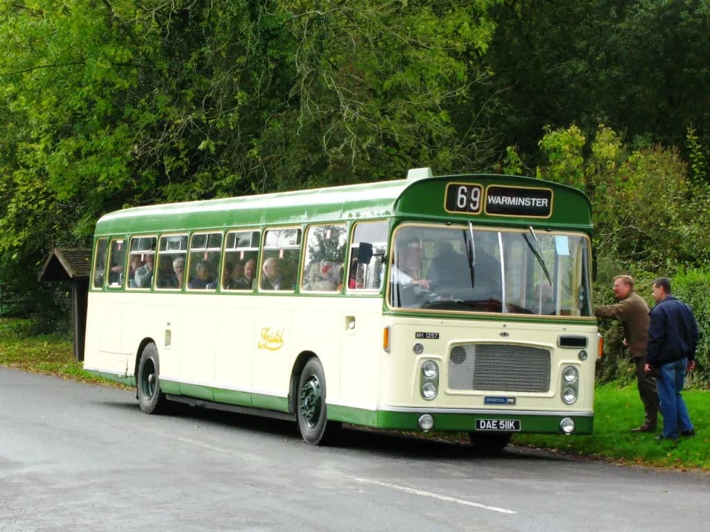A vintage cream and green bus with passengers visible through the windows is stopped on a paved road beside a grassy bank, while two men stand near the front of the bus.